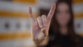 Sports gif. Sierra Sass, a member of the Loyola Ramblers Softball team, holds up a rock on symbol. The camera starts at the symbol then pans towards her face, which comes into focus and she smiles at us.
