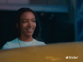 Sponsored gif.  Pink bubble letter text that reads "Blushing" flies into frame as a woman looks away from it with a smile, a shy or bashful expression on her face. The Tinder logo appears in the bottom right corner.