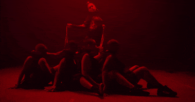 Music video gif. From the video for Bend and Snap, lit by red lights and dressed like a dominatrix, Dizzy Fae holds onto four dancers with leashes as they sit with legs around each other, leaning back.