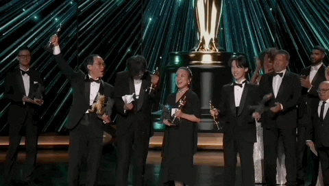 Oscars 2024 gif. The Godzilla Minus One team wins Visual Effects. They happily raise their arms and wave their trophies in the air while award presenters applaud behind them. 