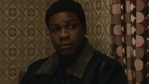 TV gif. John Boyega as Leroy in Small Axe: Red, White, and Blue tilts his head and purses his lips with a sigh as he lifts a glass to cheers.