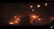 video game gif. A futuristic caped warrior, viewed from behind, thrusts his fist into the air as a giant explosion occurs on a giant spacecraft far-off in the distance.