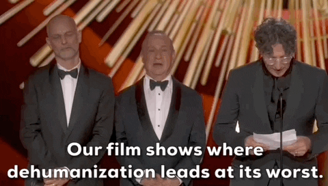 Oscars 2024 gif. Jonathan Glazer wins Best International Film for Zone of Interest. His hands tremble as he reads from a paper onstage and says, "Our film shows where dehumanization is at its worst." Two men stand upright behind him with their hands clasped in front of them.