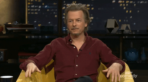 Celebrity gif. David Spade throws up his hands in a shrug as he frowns uncertainly.