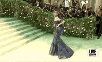 Met Gala 2024 gif. Zendaya poses in place, giving a full view of her her midnight blue and dark teal Maison Margiela tulle gown with a trumpet skirt covered in royal blue and emerald green diagonal stripes and fruit-like embellishments adorning one side of the waist and arm. 