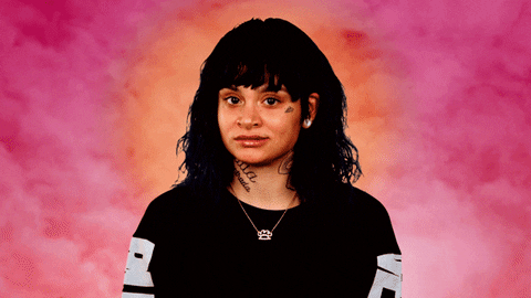 Celebrity gif. Kehlani stands in a black outfit in front of a mystical, cloudy background that has a radial gradient from orange to bright pink. She smacks on her gum while she widens her eyes suspiciously at us and looks away.