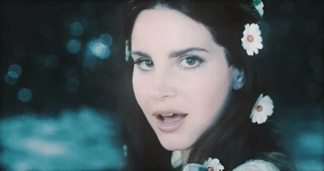 Music video gif. With daisies in her hair, Lana Del Rey winks at us over her shoulder.
