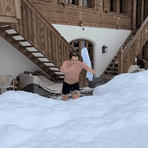 Video gif. A shirtless man dives into a fresh pile of snow and pretends to swim through it. 