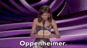 Oscars 2024 gif. Zendaya wears a glimmering, purple holographic dress that coordinates with the royal purple backdrop. She reads off of the envelope and announces Oppenheimer for Cinematography. 
