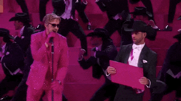 Oscars 2024 GIF. Ryan Gosling is in the middle of his performance for I'm Just Ken and he punches and breaks a wooden panel, yelling out with vigor. Background dancers lean to the right behind him and they're all wearing black cowboy hats and tuxedos.  