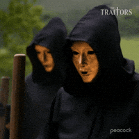 Sponsored gif. Two ominous figures in black cloaks and gold full-face masks from the Traitors rhythmically tap wooden staffs against the ground.