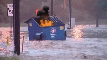 Video gif. A dumpster floats down a flooded street, containing a superimposed fire.