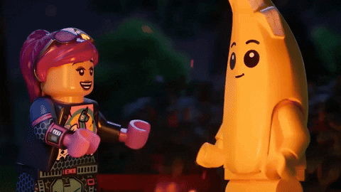 Video game gif. Clip of two Lego characters from "Lego Fortnite," one pink with a black outfit and one as a yellow banana, dance around a fire at night. The frame changes to a zoomed in view of them doing high five with their Lego hands. 