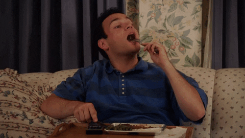 TV gif. Troy Gentile as Barry from The Goldbergs gleefully eats a forkful of a TV dinner while keeping one finger on a remote control.