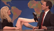 SNL gif. Kristen Wiig, dressed as a flirtatious character, legs wide above her head, uses a wheeled office chair to shuffle her hips into Seth Meyers' personal space, who cringes and covers his smile with embarrassment and discomfort.