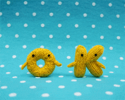 Video gif. Against a polka dot background, a knitted yellow O and K, each with tiny black eyes and hands jump up and give each other a high-five.