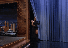 Tonight Show gif. Anitta walks onto the stage in a black dress and boots and waves with both her hands.