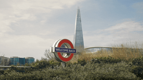 Video gif. London Tube sign reads “Welcome Back” and is surrounded by green bushes, the London Shard building towering in the background.
