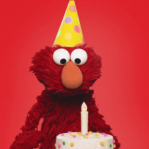 gif of Elmo on a red background wearing a party hat. A white birthday cake with a single candle sits in front of him. He smiles and laughs.