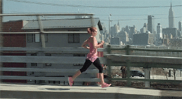 Video gif. A woman in pink runs for exercise on a elevated road as a city skyline passes in the background. 
