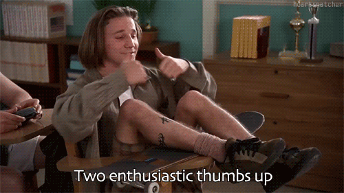 Movie gif. Breckin Meyer as Travis in Clueless sits at a small school desk with his skateboard on the desk and his legs laying over top of his skateboard. He looks up with a small and puts two thumbs up as he says, “Two enthusiastic thumbs up.”