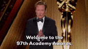 Conan O’Brien, wearing a tuxedo and looking earnest with his hands together, stands in front of a golden toned set with the Oscar statuette. Text below reads, “Welcome to the 97th Academy Awards.”