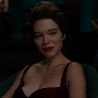 Movie gif. Léa Seydoux as Caprice in Crimes of the Future sits in the shadows as a smile spreads across her face and erupts in a giggle.