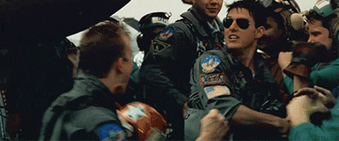Movie gif. Tom Cruise as Pete Mitchell in Top Gun screams in excitement and hugs another pilot, lifting him up in the air. Other pilots cheer around them.