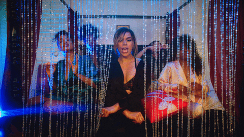 Music video gif. Dinah Jane in Heard It All Before sits at the foot of a bed, dancing in unison with her background dancers behind a beaded curtain. 