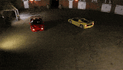 Video gif. Two red and yellow Ferrari F50s face off doing burnouts in an empty lot at night.