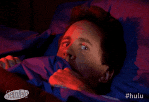 Seinfeld gif. Jerry Seinfeld as himself lies terrified in bed as he holds the blankets up tight to his face. He glances worried around the room.