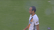 Sports gif. Landon Donovan shakes hands and hugs other soccer players on the field.