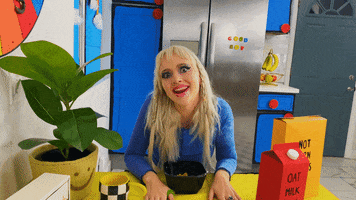 Music video gif. In the video for the song "Dumb Dumb," Mazie sits at a cartoonishly bright kitchen table and cocks her head back and forth, looking at us and singing, "la la la."
