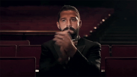 Celebrity gif. Shia Labeouf wears a tux in a dark theater and claps forcefully with an austere expression on his face.