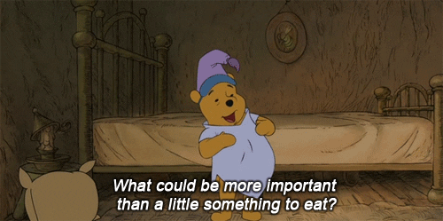 Disney gif. Winnie the Pooh stands in his all-brown bedroom, dressed in vibrant indigo pajamas with a purple nightcap and a dark blue sleeping mask. He pats the top of his belly hungrily and says, "What could be more important than a little something to eat?," which appears as text.