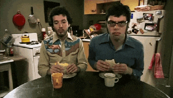 TV gif. Bret McKenzie and Jemaine Clement in Flight of the Conchords  sway rhythmically as they sit at a table eating sandwiches.