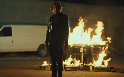 Music video gif. In his video for Selfish, Kid LAROI gazes downward dejectedly as he stands in a parking lot with a large object engulfed in flames behind him. 