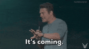 Video gif. On the 2024 Summer Game Fest stage, presenter Jason Blum, wearing a light-teal colored t-shirt, says, 'It's coming.' into the microphone he holds with his right hand. The caption at the bottom of the gif reads the same. His left hand is stretched out in a "hold on" gesture. 