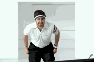Celebrity gif. Jack Gyllenhaal has a white polo and a sweatband on his head. With a  serious expression on his face, he rips his pants off to reveal short shorts. 