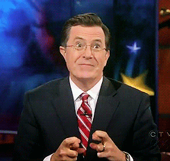TV gif. Stephen Colbert as host of the Late Show raises his eyebrows and smiles wide, crossing his fingers in a tense, awkward expression.