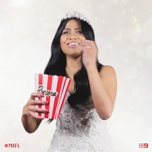 Reality TV gif. A contestant from Married at First Sight is wearing a tiara, wedding dress, and holds a box of popcorn. She eats the popcorn clumsily and eagerly and the popcorn falls everywhere as she laughs.