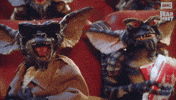 Sponsored gif. Two gremlins vibrate with excitement while sitting in movie theater seats. In the upper right corner is the AMC Fear Fest logo.
