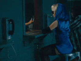 Music video gif from Sabrina Carpenter's music video for "Please Please Please." She wears a blue dress with a hood around her head, dark sunglasses, and elbow length black gloves. She is visiting Barry Keoghan in jail. He is behind glass in an orange jumpsuit. They both pick up a telephone and talk.