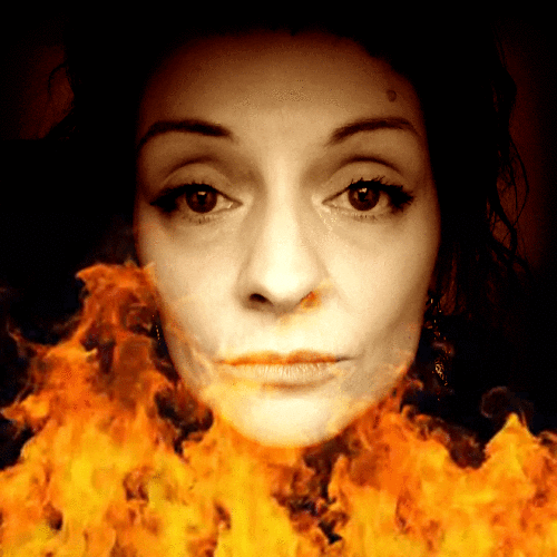 Photo gif. A woman looks at us with a blank expression on her face. Flames rise below her chin and light up her face. 