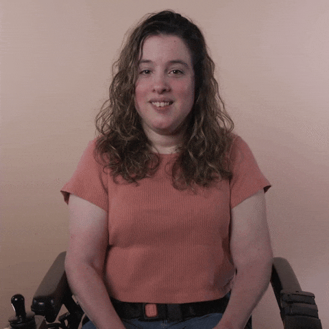 Reaction gif. A Disabled white-presenting Latina woman with cerebral palsy and kinky-curly brown hair, seated in her motorized wheelchair, hugs herself, smiles, and leans in with an impish sweetness.