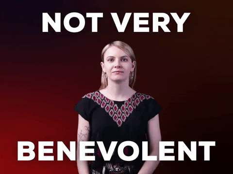 Video gif. Woman leans in and looks at us with an intense side eye, wagging a finger at the camera with a disapproving look against a dark red background. Text, "Not very benevolent." 