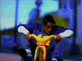 Celebrity gif. Different angles of Coolio, wearing a blue sports jersey as he rides down a street lined with palm trees on a plastic yellow tricycle.