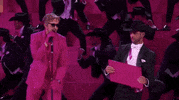 Oscars 2024 GIF. Ryan Gosling is in the middle of his performance for I'm Just Ken and he punches and breaks a wooden panel, yelling out with vigor. Background dancers lean to the right behind him and they're all wearing black cowboy hats and tuxedos.  