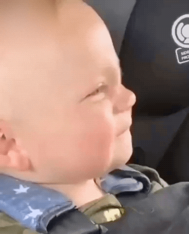 Video gif. Closeup of a baby sitting in a car seat with their eyes closed, bobbing their head to a beat, and pursing their lips together like they are singing in a cool, intense way. 