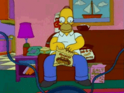 The Simpsons gif. Homer sits on a couch surrounded by junk food as his hands frantically reach into each bag and he crams the food into his mouth. 